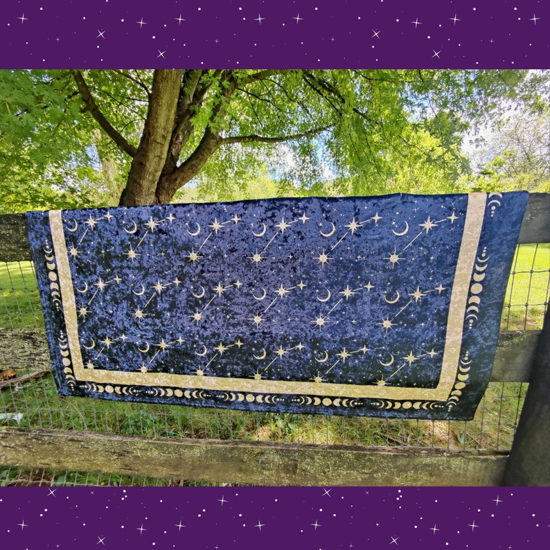 May include: A dark blue velvet tapestry with a celestial design of stars, moons, and shooting stars. The tapestry has a gold border with moon phases. The fabric is draped over a wooden fence, with a green background.