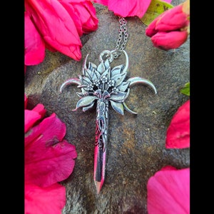 May include: A silver pendant necklace featuring a stylized floral design with a pointed tip. The pendant is shaped like a flower with a central stem and multiple petals. The pendant is surrounded by a silver chain.