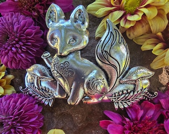 Könnte beinhalten: Eine silberne Fuchsfigur mit filigranen Details, die zwischen Pilzen und Blättern sitzt. Der Fuchs hat einen verspielten, fast cartoonartigen Ausdruck mit großen Augen und einer leicht nach oben gerichteten Schnauze. Die Figur ist von verschiedenen Blumen umgeben, darunter lila, gelbe und rote.