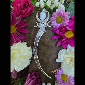 May include: A silver-toned, crescent-shaped hair accessory with a central opal-like stone, wings, and moon details. The accessory is surrounded by vibrant pink and white flowers, set against a dark, textured background.