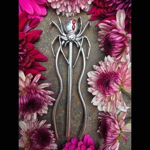May include: A silver-toned metal hair pin shaped like a spider with long, curved legs extending downward. The spider body features two small, bright red oval accents on its back. The pin is surrounded by vibrant pink and purple chrysanthemum flowers.
