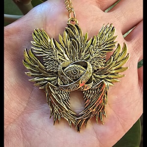 Może przedstawiać: Złoty naszyjnik z wisiorkiem o szczegółowym wzorze. Wisiorek przedstawia centralny motyw oka w pierścieniu, otoczony stylizowanymi skrzydłami. Naszyjnik ma delikatny łańcuszek.