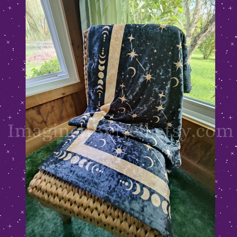 May include: A dark blue blanket with a celestial design of moons, stars, and constellations in a gold color. The blanket is draped over a wicker chair, creating a cozy and inviting scene. The blanket's design is repeated on the border.