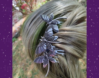 May include: A silver hair comb with three dragonfly motifs. The dragonfly wings have etched detailing. The comb is set in blonde hair, with a blurred outdoor backdrop.