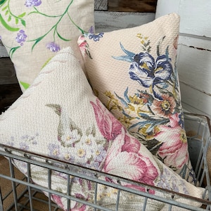 May include: Three floral patterned throw pillows in a wire basket. The pillows are in shades of pink, green, and blue. The pillows are made of a soft, textured fabric.