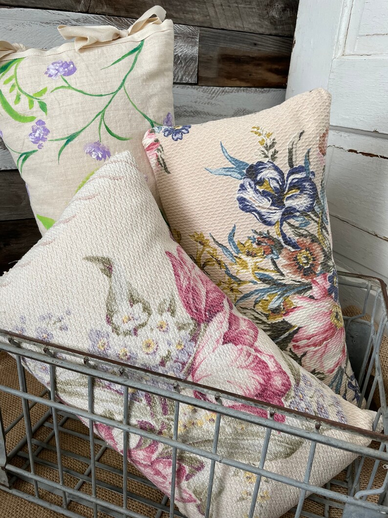 May include: Three floral patterned throw pillows in a wire basket. The pillows are in shades of pink, green, and blue. The pillows are made of a soft, textured fabric.
