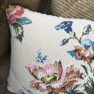 May include: A beige pillow with a floral pattern featuring pink, blue, and brown flowers. The pillow has a textured surface.