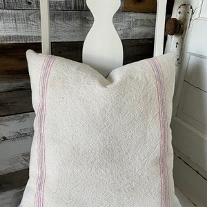 Puede incluir: Una almohada de lino blanco con rayas rojas y azules en los bordes. La almohada está sentada en una silla blanca con un asiento a rayas.