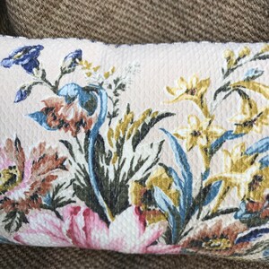 May include: A cream-colored pillow with a floral pattern featuring pink, blue, yellow, and brown flowers. The pillow has a textured surface.