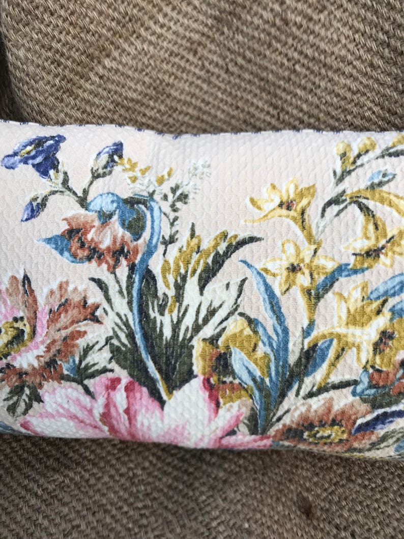 May include: A cream-colored pillow with a floral pattern featuring pink, blue, yellow, and brown flowers. The pillow has a textured surface.
