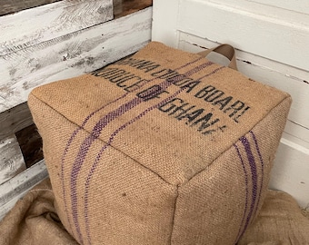 May include: A cube-shaped burlap ottoman with a tan color and purple stripes. The text "Ghana Cocoa Board" is printed on the side. The ottoman has a tan handle and sits on a burlap surface.