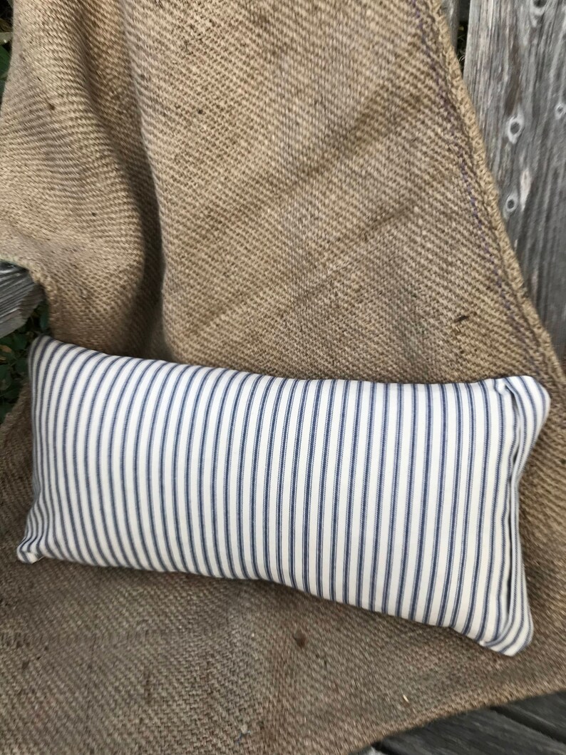 May include: A white and blue striped rectangular pillow with a subtle sheen. The pillow is sitting on a brown burlap surface.