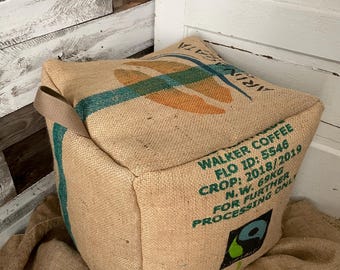 May include: A square coffee sack pillow made from burlap. The pillow has a teal and orange design on the top and the text "WALKER COFFEE" and other details printed on the side. The pillow is sitting on a burlap surface.
