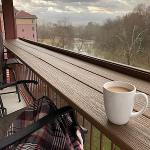 May include: A white ceramic mug filled with coffee sits on a wooden table with a plaid blanket draped over a chair. The table is on a balcony overlooking a river and trees.