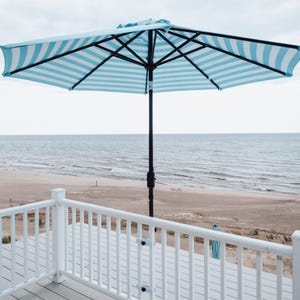 May include: A blue and white striped patio umbrella with a black metal frame, standing on a white wooden deck overlooking a sandy beach and ocean.