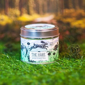 May include: A small, round candle tin with a silver lid. The tin features a teal and white design with illustrations of hares and plants. The text "THE HARE" is printed on the front, along with "HAND MADE SUSTAINABLE CANDLE". The candle sits on green grass.