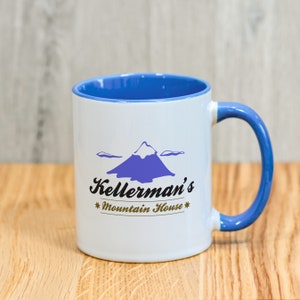 Può includere: Tazza in ceramica bianca con interno e manico blu. La tazza presenta una grafica di montagna viola e il testo "Kellerman's Mountain House" in nero e oro. Su una superficie di legno.