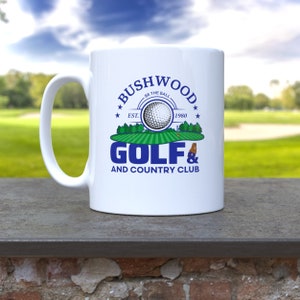 May include: White ceramic mug with a blue and green design. The design features a golf ball, a golf course, and the text "Bushwood Golf & Country Club" with the text "Be the Ball" and "Est. 1980" in smaller font.