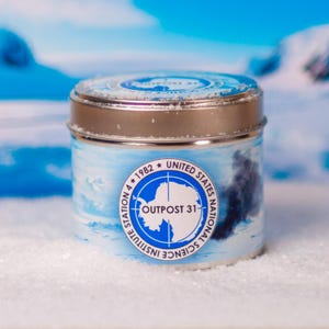 May include: A silver tin candle with a blue and white label. The label features a map of Antarctica with the text "Outpost 31" and "United States National Science Foundation 4 * 1982".
