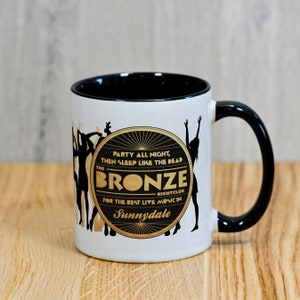 May include: White ceramic mug with a black rim and handle. The mug features a black and gold design with the text "PARTY ALL NIGHT, THEN SLEEP LIKE THE DEAD THE BRONZE NIGHTCLUB FOR THE BEST LIVE MUSIC IN Sunnydale".