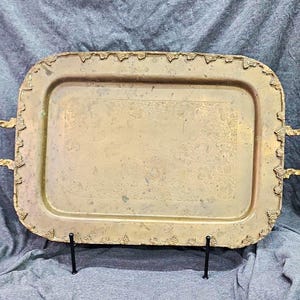 May include: A rectangular brass serving tray with a weathered patina, featuring ornate decorative handles on both sides and a detailed embossed floral pattern along the border and the central recessed area of the metal surface.