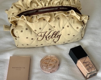 May include: A beige quilted cosmetic bag with a ruffled top and the name "Kelly" embroidered in brown. The bag is surrounded by makeup products, including a compact, a foundation bottle, and a Patrick Ta product.