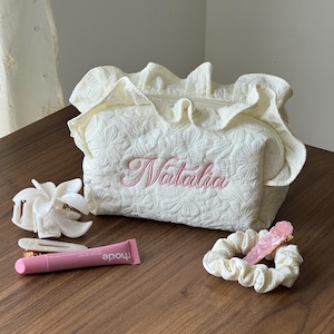 May include: Cream-colored quilted cosmetic bag with the name "Natalia" embroidered in pink. The bag has a ruffled edge and a zipper closure. Accessories include a white flower hair clip, a pink lip tint, and a matching scrunchie with a pink hair clip.