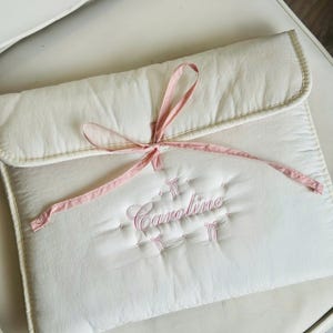 May include: Cream-colored quilted baby accessory with the name "Caroline" embroidered in pink script. The accessory is tied with a pink ribbon and has a soft, padded appearance. The item is placed on a white chair.