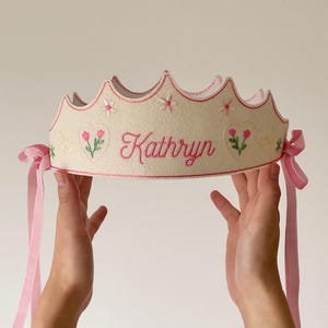 May include: An off-white felt crown featuring pink embroidered details, including the name Kathryn, heart shapes, flowers, and butterflies. The crown has a scalloped top edge with pink stitching and is secured with light pink ribbon ties on both sides.