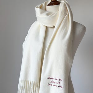 May include: Cream-coloured scarf with fringe detail, draped on a mannequin. The scarf has the embroidered text "Always love you always will love nana xoxo" in a burgundy colour. The scarf is made of a soft, woven material.