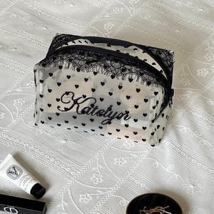 May include: A rectangular cosmetic bag made of sheer white mesh fabric featuring a pattern of small black hearts. The bag is accented with black lace trim along the top zipper and displays the name Katelyn embroidered in black script on the front.