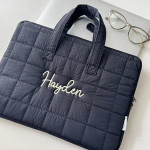 May include: A navy blue quilted laptop sleeve with a handle and the name "Hayden" embroidered in white script. The sleeve is rectangular and has a zipper closure. A pair of eyeglasses rests on a laptop in the background.