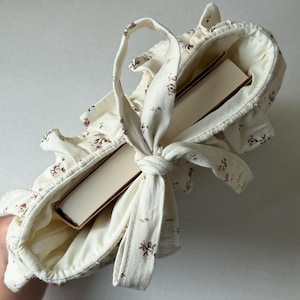Puede incluir: Un bolso tote de color crema con un estampado floral, que contiene dos libros. El bolso tiene un borde con volantes y un asa atada. El dise&ntilde;o floral presenta peque&ntilde;as flores marrones. Los libros tienen cubiertas beige.
