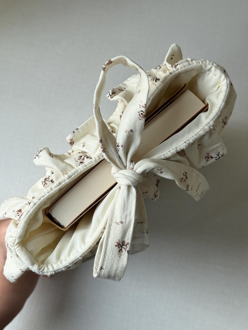 Puede incluir: Un bolso tote de color crema con un estampado floral, que contiene dos libros. El bolso tiene un borde con volantes y un asa atada. El dise&ntilde;o floral presenta peque&ntilde;as flores marrones. Los libros tienen cubiertas beige.