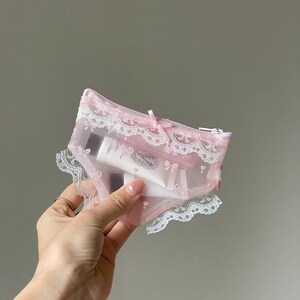 Könnte beinhalten: Ein kleines, hellrosa Netz-Etui in Form einer Unterhose, verziert mit weißer Spitzenborte an den Rändern und einer kleinen Satinschleife in der Mitte. Das halbtransparente Material gibt den Blick auf eine weiße Kosmetiktube im Inneren frei.