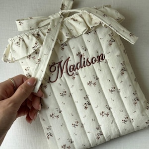 Puede incluir: Funda de tela acolchada color crema con un estampado floral y el nombre "Madison" bordado en marr&oacute;n. La funda tiene un borde superior con volantes y un cierre de lazo.
