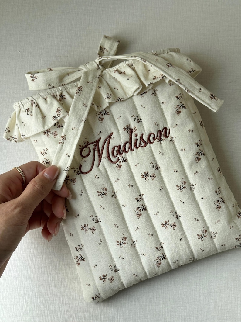 Puede incluir: Funda de tela acolchada color crema con un estampado floral y el nombre "Madison" bordado en marr&oacute;n. La funda tiene un borde superior con volantes y un cierre de lazo.
