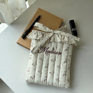 Puede incluir: Una funda acolchada para tableta de color crema con un estampado floral y el nombre "Madison" bordado en marr&oacute;n. La funda tiene una parte superior con volantes y un lazo. Un bol&iacute;grafo, un cuaderno y un l&aacute;piz labial tambi&eacute;n est&aacute;n sobre la mesa.