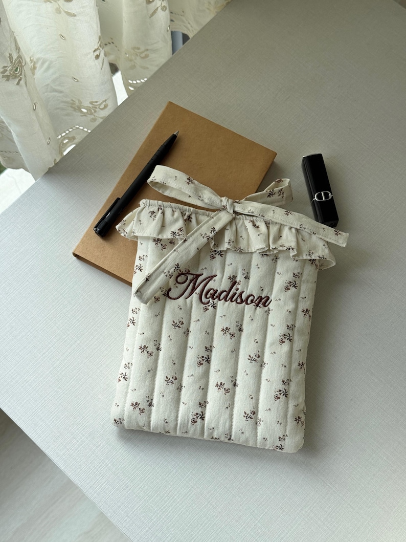 Puede incluir: Una funda acolchada para tableta de color crema con un estampado floral y el nombre "Madison" bordado en marr&oacute;n. La funda tiene una parte superior con volantes y un lazo. Un bol&iacute;grafo, un cuaderno y un l&aacute;piz labial tambi&eacute;n est&aacute;n sobre la mesa.