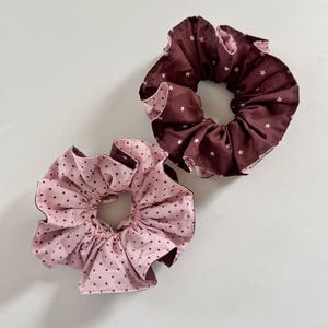 May include: Two fabric hair scrunchies. One scrunchie is dark brown with a small white star pattern. The second scrunchie is light pink with a small dark brown polka dot pattern. Both items feature gathered fabric with visible stitching along the edges.
