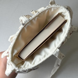 Puede incluir: Bolso tote de color crema con un estampado floral y ribete con volantes. El bolso est&aacute; abierto, revelando un libro con una cubierta marr&oacute;n. El bolso tiene dos correas y est&aacute; hecho de tela suave.