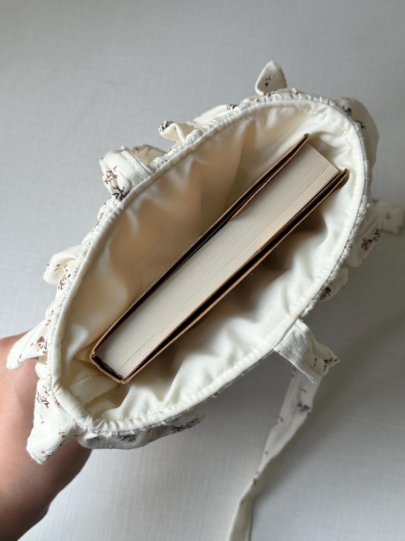 Puede incluir: Bolso tote de color crema con un estampado floral y ribete con volantes. El bolso est&aacute; abierto, revelando un libro con una cubierta marr&oacute;n. El bolso tiene dos correas y est&aacute; hecho de tela suave.