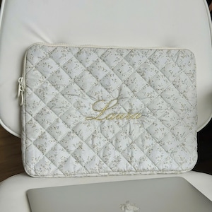 May include: White quilted laptop sleeve with a floral pattern and the name "Laura" embroidered in gold. The sleeve has a zip closure and is on a white chair. A silver laptop is below the sleeve.