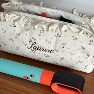 May include: A cream-colored floral print cosmetic bag with the name "Lauren" embroidered on the front. A teal and orange hair styling tool is in front of the bag. The bag has a ruffled trim and a zipper closure.