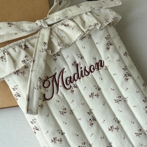 Puede incluir: Funda acolchada para port&aacute;til de color crema con un estampado floral y el nombre "Madison" bordado en escritura marr&oacute;n. La funda tiene un borde superior con volantes y un cierre de lazo. Un cuaderno marr&oacute;n y un l&aacute;piz labial negro est&aacute;n en el fondo.