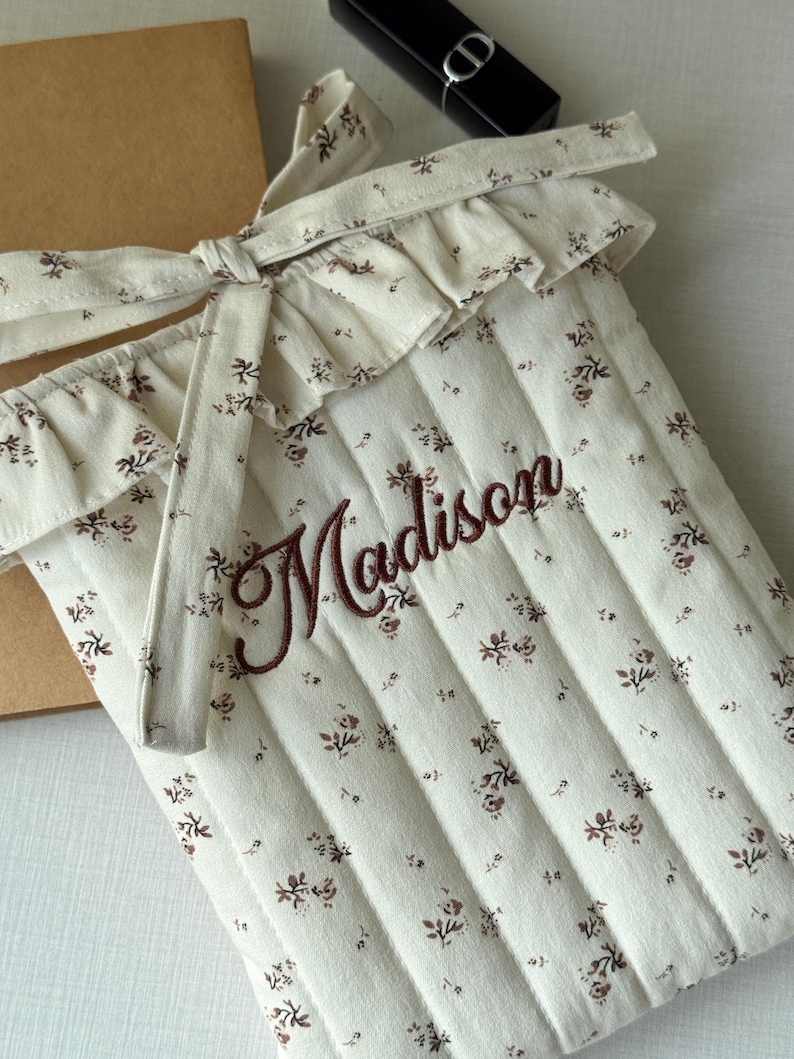 Puede incluir: Funda acolchada para port&aacute;til de color crema con un estampado floral y el nombre "Madison" bordado en escritura marr&oacute;n. La funda tiene un borde superior con volantes y un cierre de lazo. Un cuaderno marr&oacute;n y un l&aacute;piz labial negro est&aacute;n en el fondo.