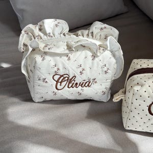 May include: Cream-colored, quilted cosmetic bag with a floral pattern and the name "Olivia" embroidered in brown. The bag has a ruffled top and a zipper closure. A smaller, similar bag with polka dots is also visible.