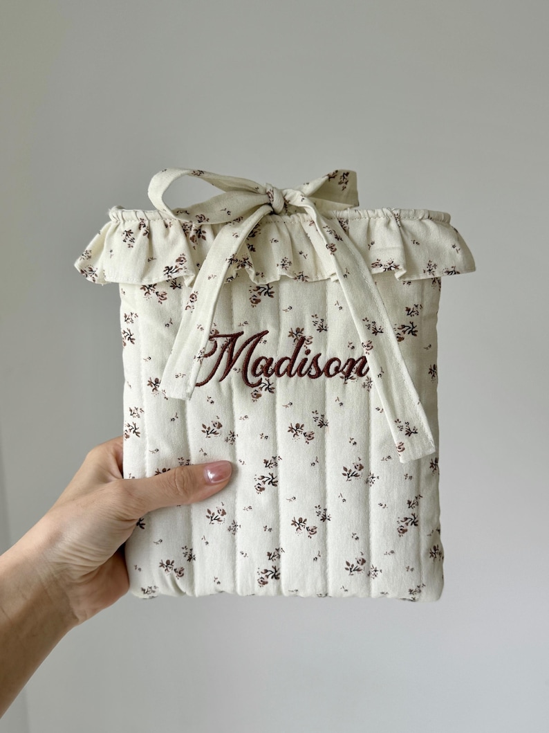 Puede incluir: Funda acolchada de color crema con un estampado floral y el nombre "Madison" bordado en marr&oacute;n. El borde superior presenta un ribete con volantes y un lazo. La funda est&aacute; dise&ntilde;ada para guardar una tableta o un lector de libros electr&oacute;nicos.