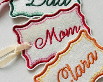 May include: Three white felt gift tags with scalloped edges and embroidered names: "Dad" in green, "Mom" in red, and "Clara" in orange. Each tag has a matching ribbon for attaching to gifts. A charming and personalized gift accessory.