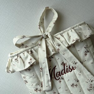 Puede incluir: Tela acolchada de color crema con un estampado floral y el nombre "Madison" bordado en marr&oacute;n. El borde superior tiene un ribete con volantes y un lazo de tela a juego atado en la parte superior. El dise&ntilde;o general es suave y delicado.